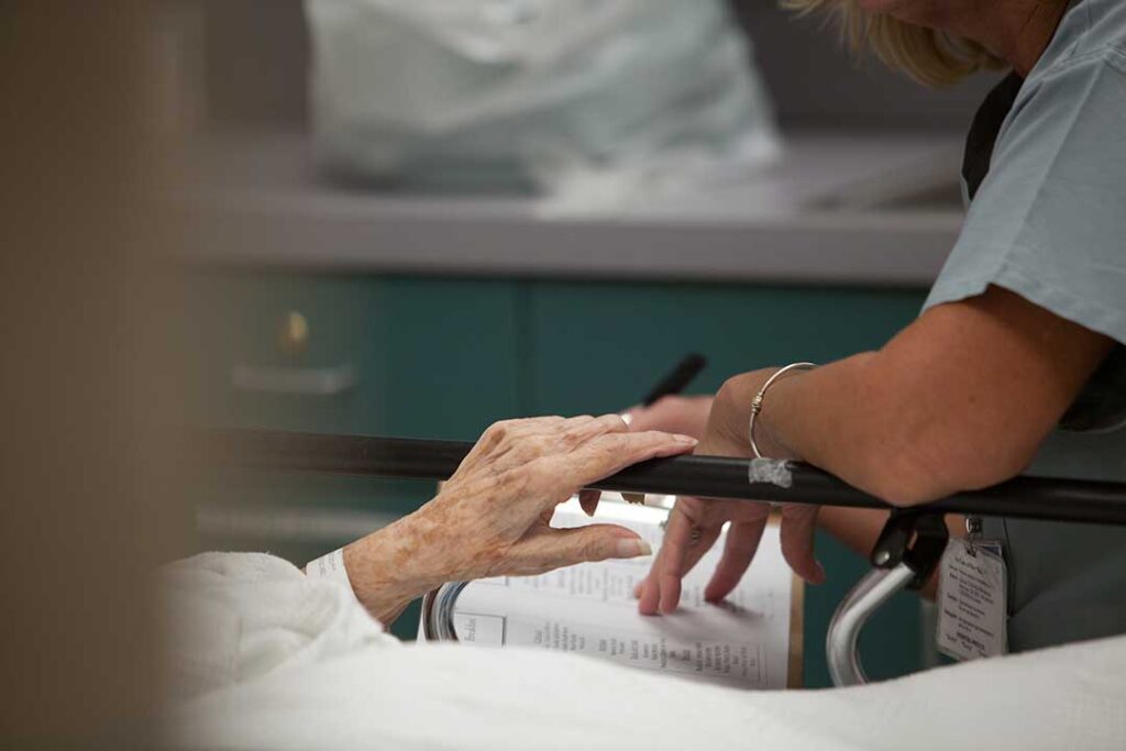 Older adults share a meal while a caregiver assists a resident in a bright community dining room.